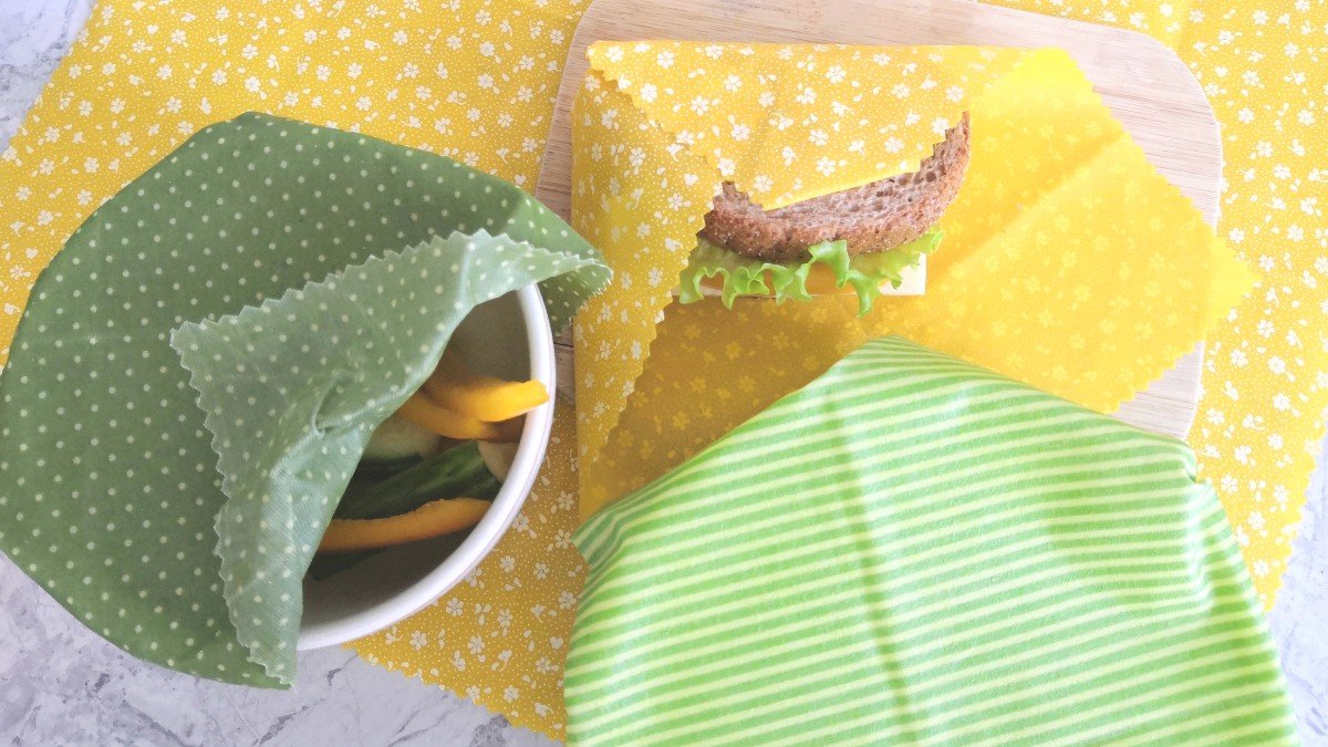 Sandwich, cucumber, bell pepper and a bowl wrapped in yellow and green beeswax wraps on a marble counter