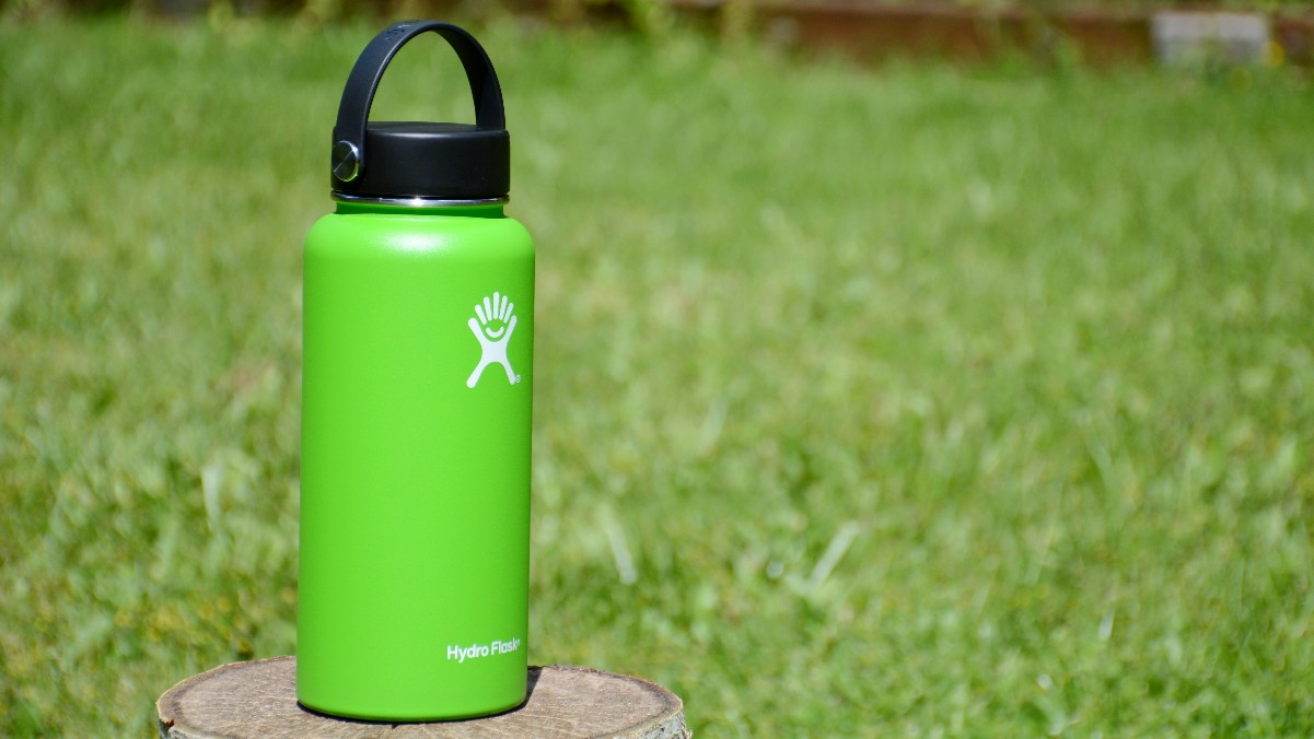 a green Hydro Flask bottle standing on a block of wood with green grass behind it